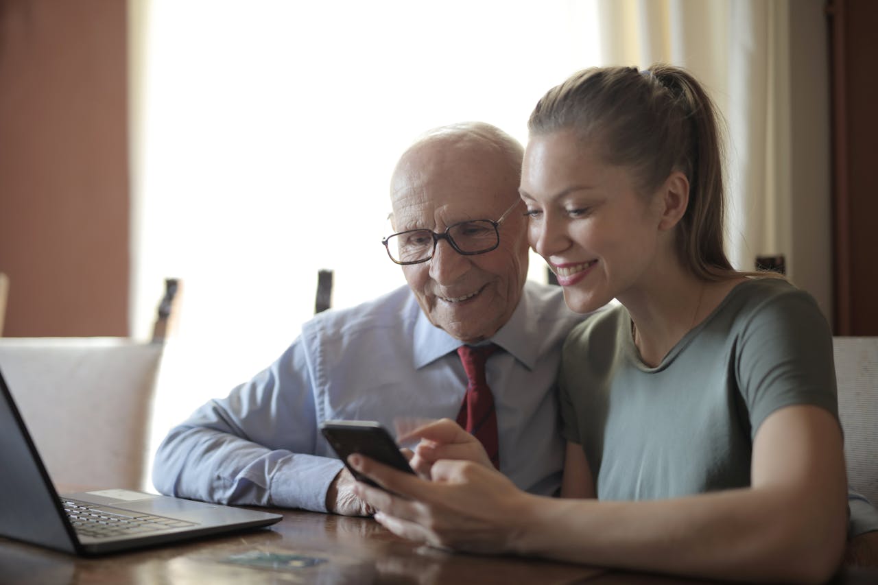 About Smiling young woman in casual clothes showing smartphone to interested senior grandfather in formal shirt and eyeglasses while sitting at table near laptop