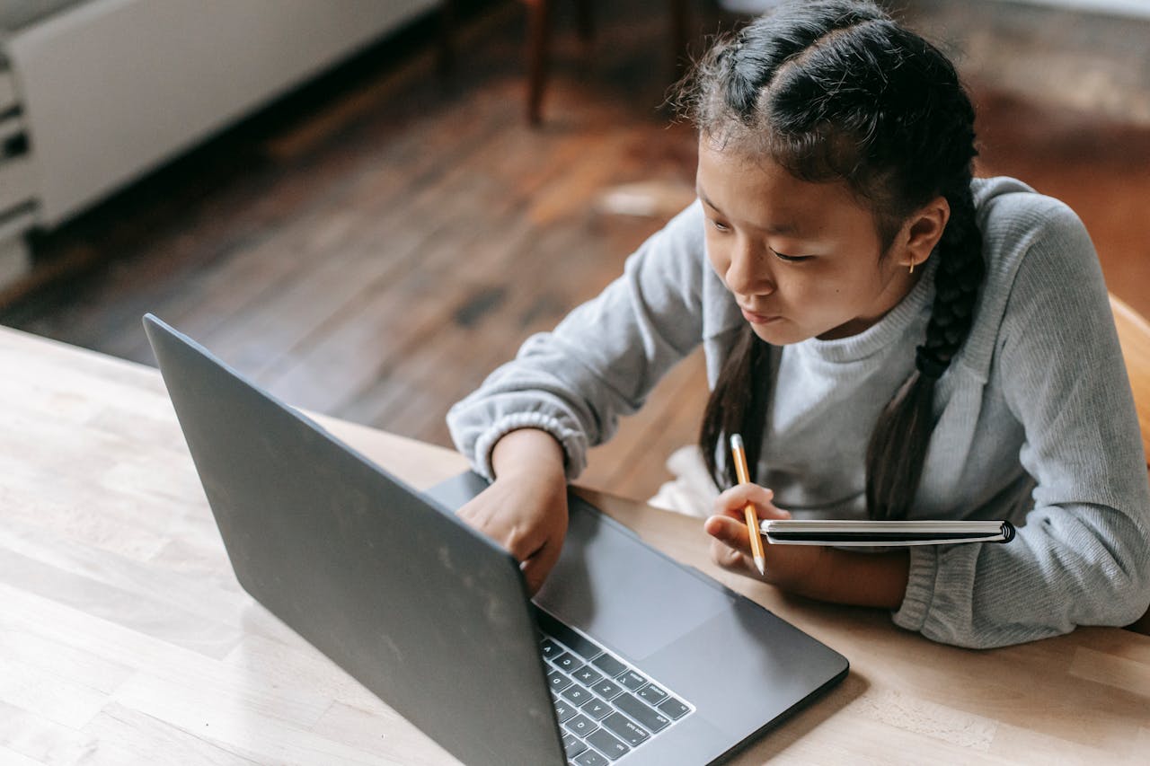 Services From above of concentrated Asian schoolgirl with pigtails sitting at wooden table with copybook and pencil and browsing laptop for school task
